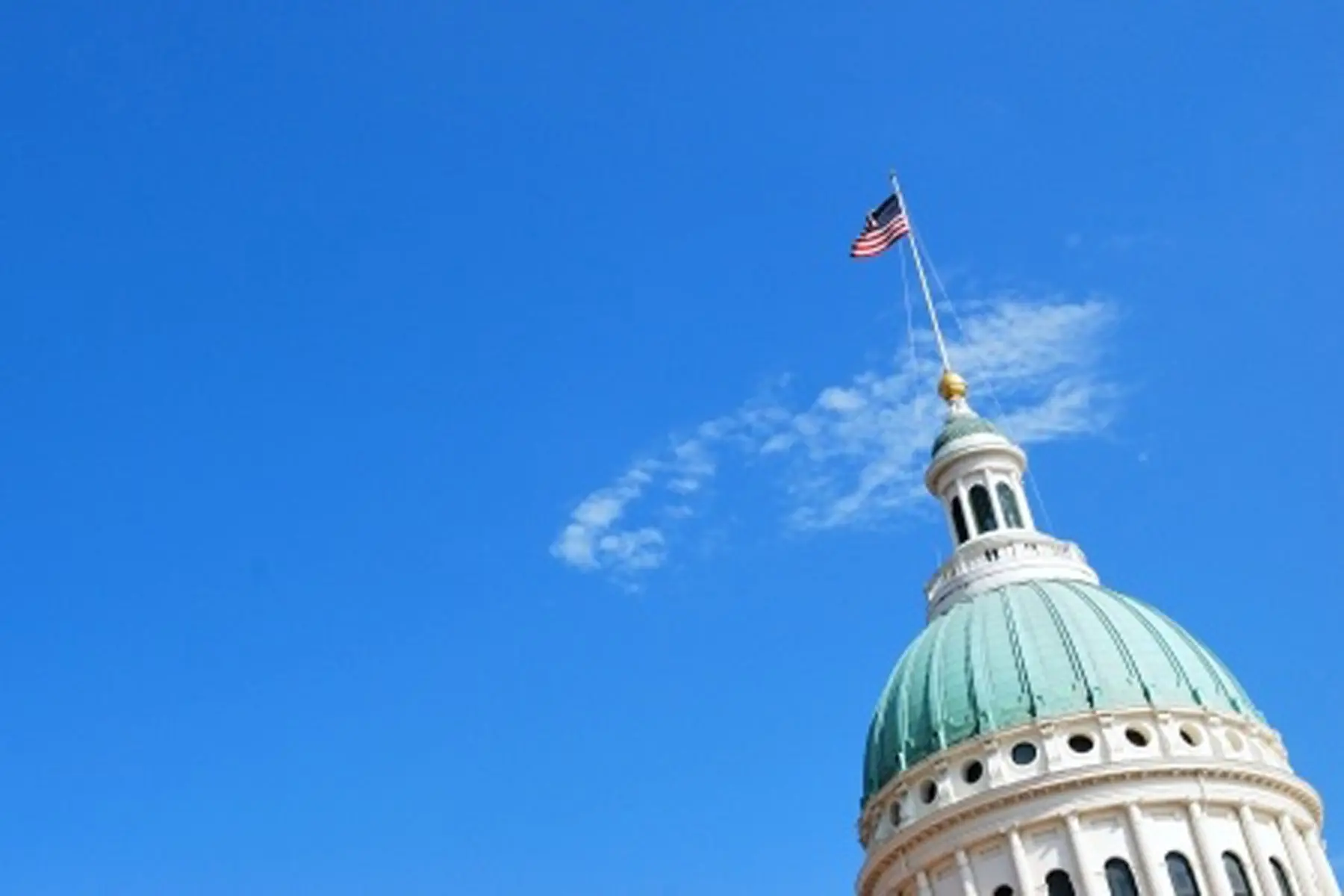 The dome of a government building with a green roof and decorative lantern under a clear blue sky, featuring an American flag on top.